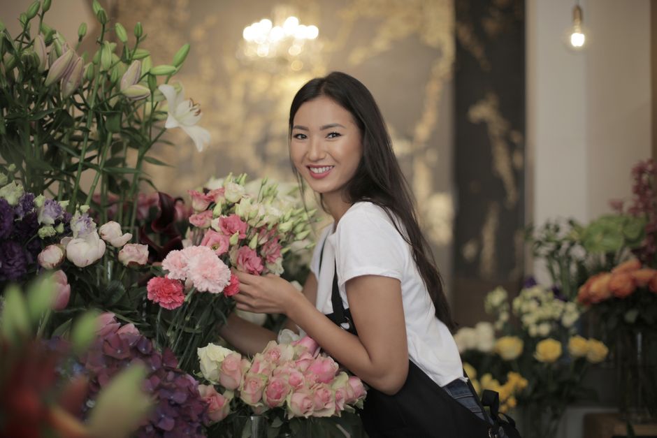 A young woman with long dark hair and a cheerful expression is holding a bouquet of pink and white carnations in a florist shop. She is wearing a white t-shirt and a black apron, standing amidst an ar