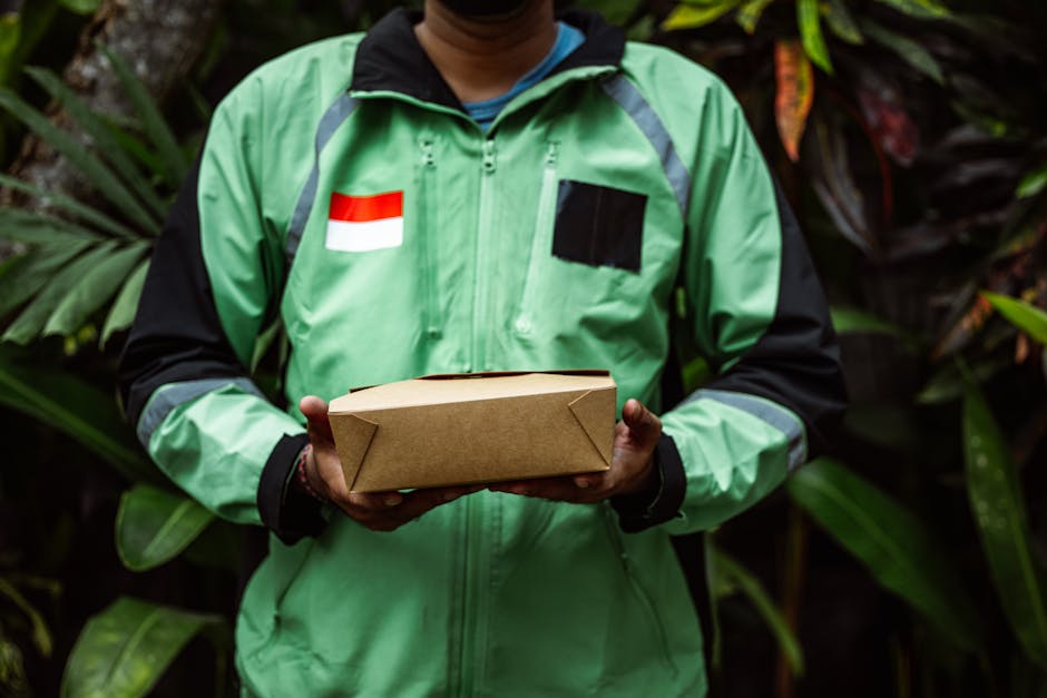A person wearing a green and black jacket with a red and white flag patch is holding a rectangular cardboard package, standing outdoors amidst lush green foliage with large leaves. The individual’s 