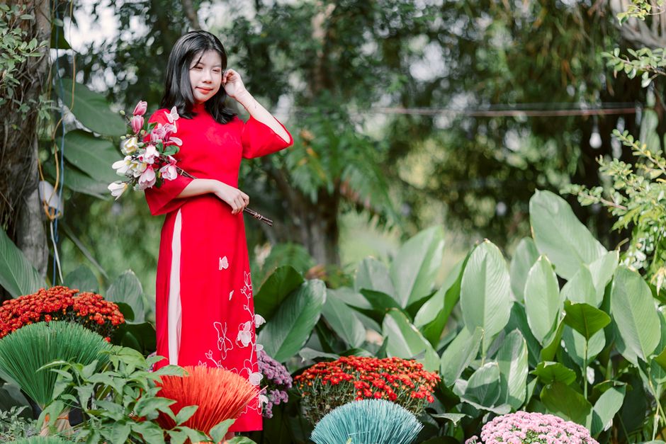 A young woman dressed in a vibrant red áo dài holds a bouquet of fresh, colorful flowers including pink orchids and white blossoms, with green foliage. She stands amidst lush, large-leafed tropical 