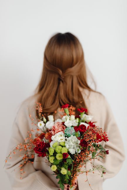 A person with shoulder-length brown hair, dressed in a beige sweater, is holding a large, vibrant floral arrangement behind their back. The bouquet features a variety of fresh flowers including red ro