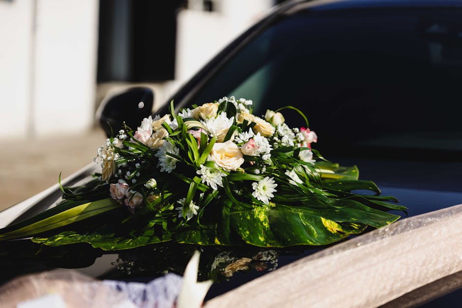 A fresh floral bouquet arrangement resting on the hood of a car, featuring white roses, daisy-like white flowers, and small pink accents, accented with lush green foliage and wrapped in clear cellopha
